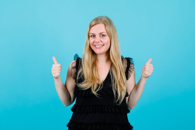 Glimlachend blond meisje kijkt naar de camera door perfecte gebaren op een blauwe achtergrond te tonen