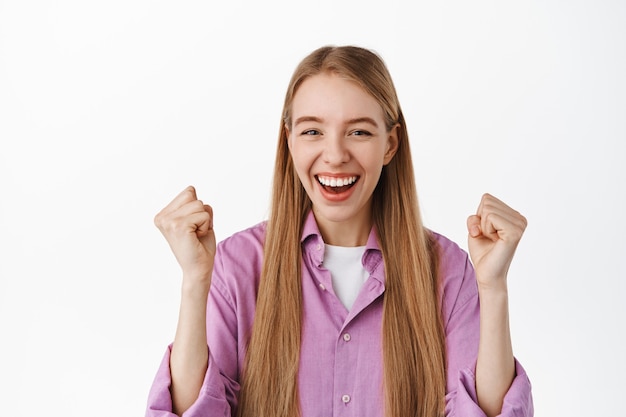 Gratis foto glimlachend blond meisje viert succes, bereikt doel, zegt ja en steekt vuisten op van vreugde, triomfeert, staande over een witte muur