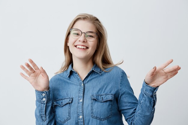 Glimlachend breed positief wijfje met blond steil haar, dat denimoverhemd draagt, dat tegen grijze blinde muur stelt. Gelukkig studentenmeisje dat positieve emoties toont na het ontvangen van goed teken