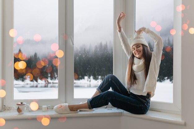 Glimlachend gelukkig jonge aantrekkelijke vrouw in stijlvolle witte gebreide trui, sjaal en muts om thuis te zitten op de vensterbank met Kerstmis plezier hand in hand omhoog, winter bos achtergrond weergave, lichten bokeh