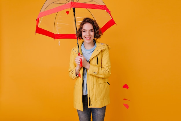 Glimlachend gelukkig Kaukasisch meisje dat rode parasol op gele muur houdt. Binnenfoto van bevallige jonge vrouw met golvend haar die zich onder paraplu bevinden.
