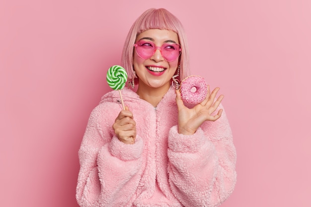 Glimlachend glamour Aziatische tienermeisje kijkt graag opzij houdt lolly op stok en smakelijke donut