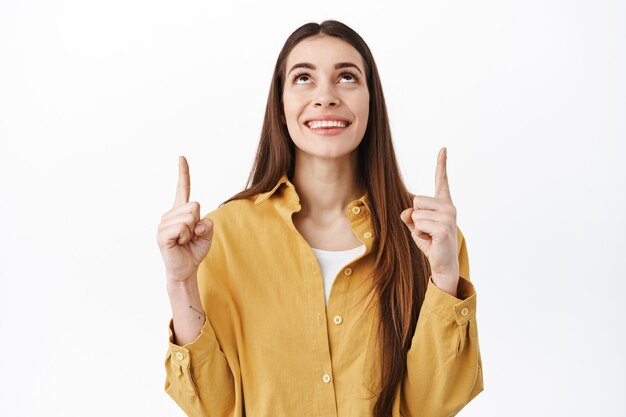 Glimlachend hoopvol meisje dat er dromerig uitziet, omhoog wijst en de tekenpromo hierboven leest, topadvertentie of logo toont, over een witte muur staat in casual stijlvolle kleding