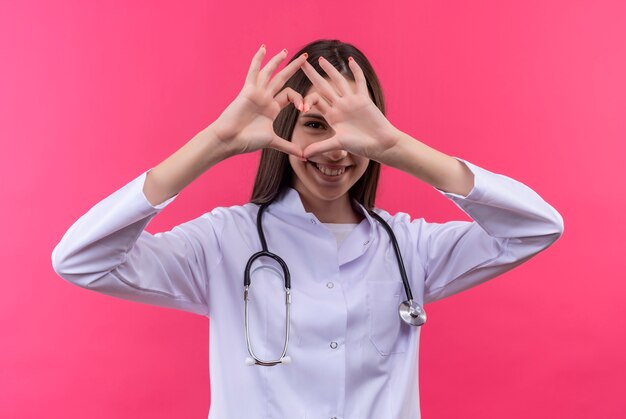 Glimlachend jong artsenmeisje dat stethoscoop medische toga draagt die hartgebaar op geïsoleerde roze achtergrond toont