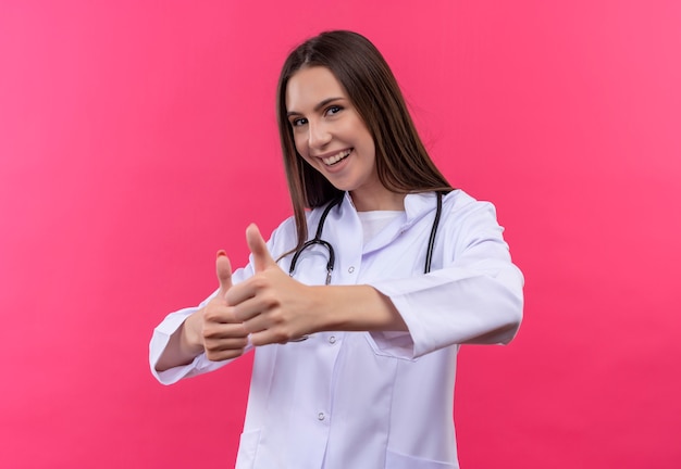 Glimlachend jong artsenmeisje die stethoscoop medische toga dragen haar duimen omhoog op geïsoleerde roze achtergrond