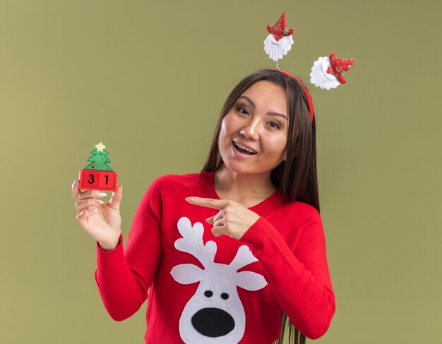 Glimlachend jong Aziatisch meisje dat de hoepelholding van het Kerstmishaar en punten op Kerstmisstuk speelgoed draagt dat op olijfgroene achtergrond wordt geïsoleerd