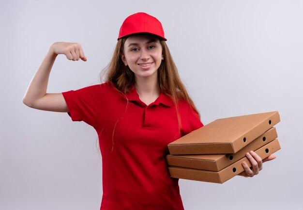 Glimlachend jong bezorgmeisje in de rode uniforme pakketten van de holdingspizza die sterk gebaar op geïsoleerde witte muur doen