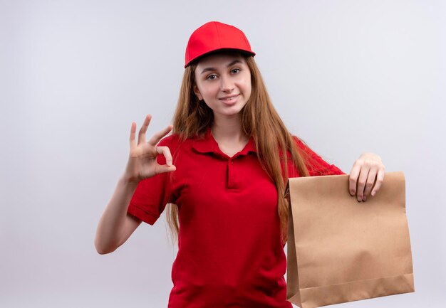 Glimlachend jong bezorgmeisje in rood uniform doet ok teken en houdt papieren zak op geïsoleerde witte ruimte