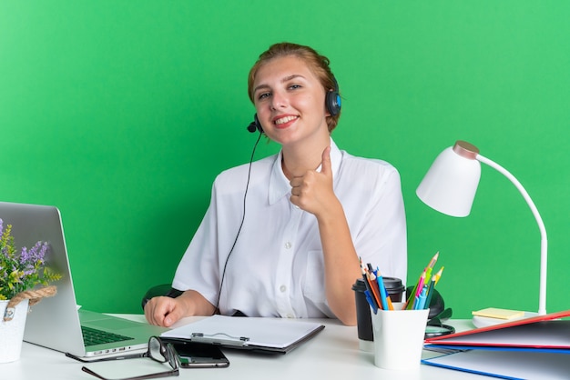 Gratis foto glimlachend jong blond callcentermeisje met een hoofdtelefoon die aan een bureau zit met uitrustingsstukken die naar een camera kijken die duim omhoog laat zien op een groene muur