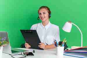 Gratis foto glimlachend jong blond callcentermeisje met hoofdtelefoon zittend aan een bureau met uitrustingsstukken met klembord kijkend naar camera geïsoleerd op groene muur