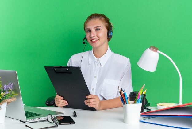 Glimlachend jong blond callcentermeisje met hoofdtelefoon zittend aan een bureau met uitrustingsstukken met klembord kijkend naar camera geïsoleerd op groene muur