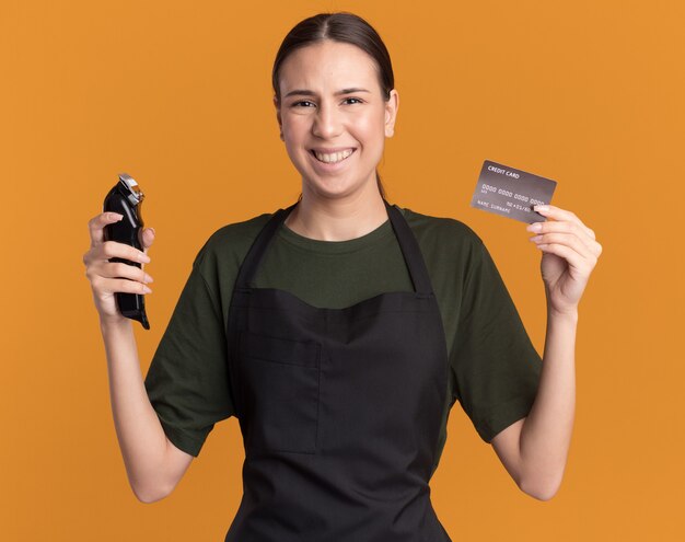 Glimlachend jong brunette kappersmeisje in uniform houdt tondeuses en creditcard geïsoleerd op een oranje muur met kopieerruimte