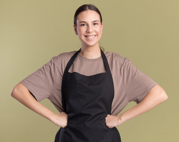 Glimlachend jong brunette kappersmeisje in uniform zet vuisten op taille