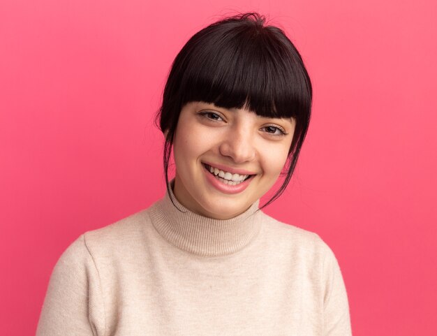 Glimlachend jong brunette kaukasisch meisje kijkt naar camera geïsoleerd op roze muur met kopieerruimte