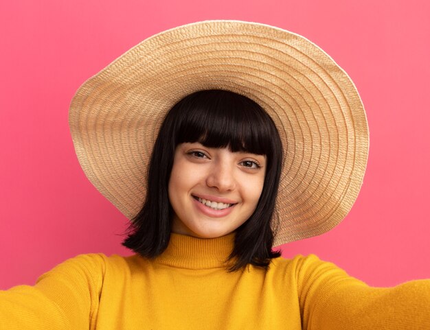 Glimlachend jong brunette kaukasisch meisje met strandhoed doet alsof ze een camera vasthoudt die selfie neemt geïsoleerd op roze muur met kopieerruimte