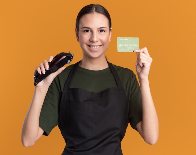 Glimlachend jong kappermeisje in eenvormige tondeuse en creditcard van de holdingshaar op oranje