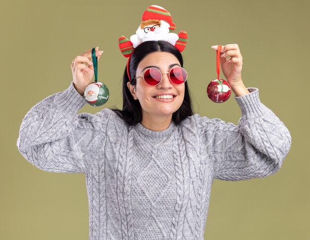 Glimlachend jong Kaukasisch meisje dat de hoofdband van de Kerstman draagt met een bril met kerstballen in de buurt van het hoofd geïsoleerd op een olijfgroene muur