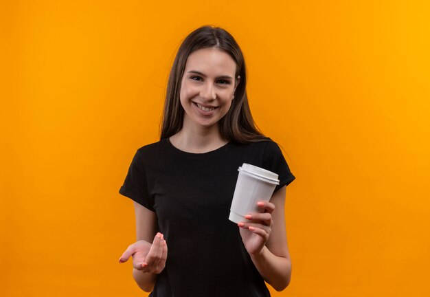Glimlachend jong Kaukasisch meisje dat een zwarte kop van de t-shirtholding van koffie draagt wijst naar camera op geïsoleerde oranje achtergrond