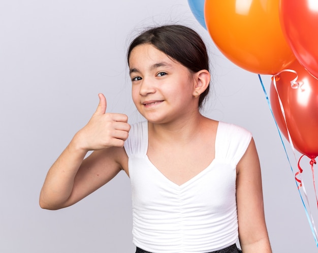 Glimlachend jong kaukasisch meisje dat met heliumballonnen staat en omhoog duimt geïsoleerd op een witte muur met kopieerruimte