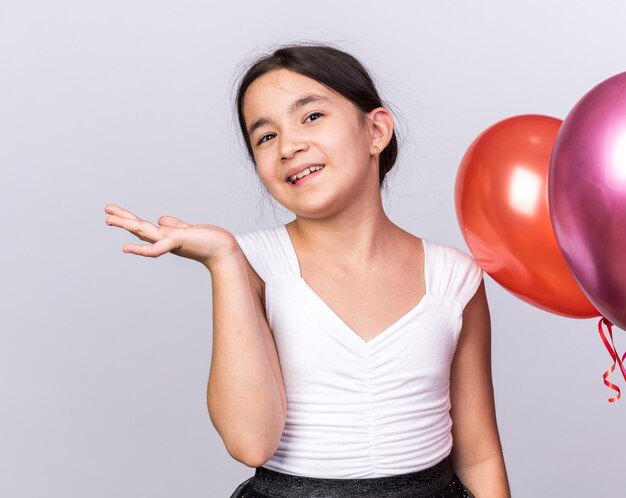 glimlachend jong kaukasisch meisje dat zich met heliumballonnen bevindt die hand open houden geïsoleerd op witte muur met exemplaarruimte
