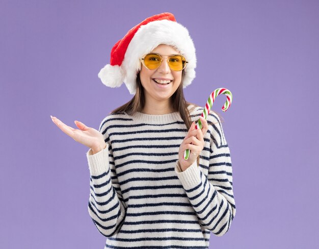 Glimlachend jong kaukasisch meisje in zonnebril met kerstmuts met snoepgoed geïsoleerd op paarse muur met kopieerruimte