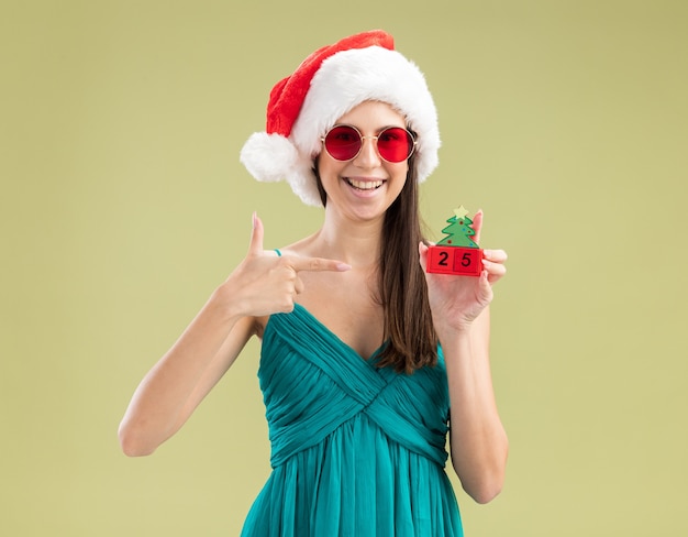 Glimlachend jong kaukasisch meisje in zonnebril met santahoed die en op het ornament van de kerstmisboom houden richten