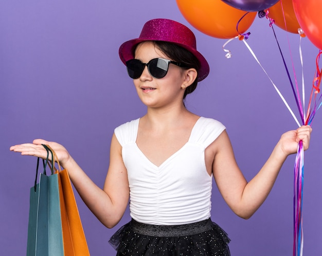 Glimlachend jong kaukasisch meisje in zonnebril met violet feestmuts met heliumballonnen en boodschappentassen kijkend naar kant geïsoleerd op paarse muur met kopieerruimte