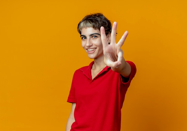 Glimlachend jong Kaukasisch meisje met pixiekapsel die drie tonen bij camera die op oranje achtergrond met exemplaarruimte wordt geïsoleerd