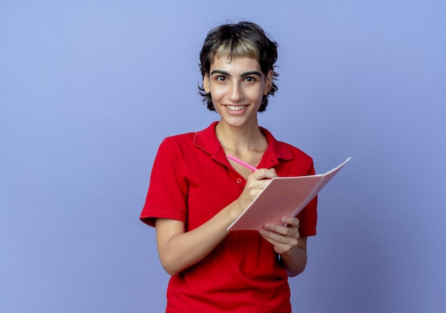 Glimlachend jong Kaukasisch meisje met pixiekapsel die pen en notitieblok houden die iets schrijven dat op purpere achtergrond met exemplaarruimte wordt geïsoleerd