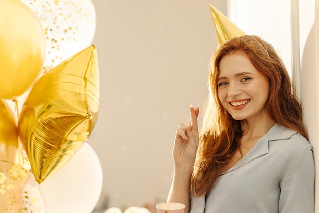 Glimlachend jong kaukasisch roodharig meisje kijkend naar camera die vingers kruist terwijl ze met ballonnen binnenshuis staat Mood lifestyle concept