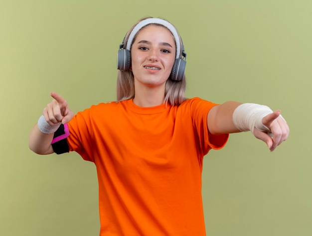 Glimlachend jong kaukasisch sportief meisje met beugels op koptelefoon met hoofdband en polsbandjes en telefoonarmband wijst op camera met twee handen