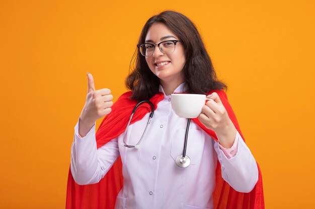 Glimlachend jong kaukasisch superheldmeisje met doktersuniform en stethoscoop met een bril met kopje thee met duim omhoog geïsoleerd op de muur