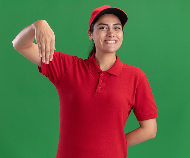 Glimlachend jong leveringsmeisje die eenvormig en GLB dragen die iets houden dat op groene muur wordt geïsoleerd beweren