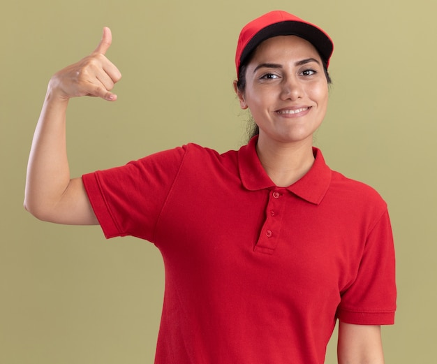 Glimlachend jong leveringsmeisje die eenvormig met GLB dragen die duim tonen die omhoog op olijfgroene muur wordt geïsoleerd