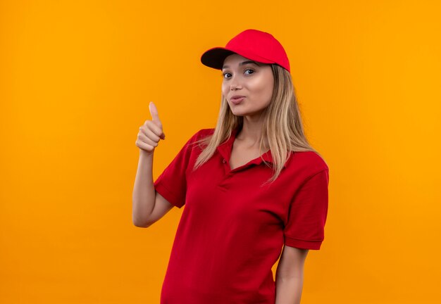 Glimlachend jong leveringsmeisje die rood uniform dragen en haar duim GLB omhoog geïsoleerd op oranje muur