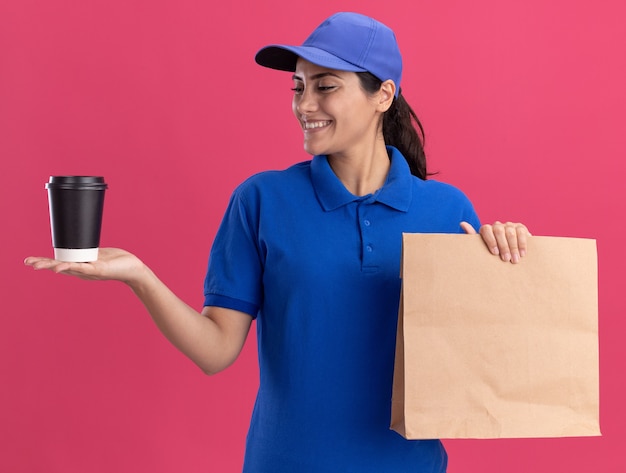 Glimlachend jong leveringsmeisje die uniform met GLB dragen die document voedselpakket houden en kop van koffie in haar hand bekijken die op roze muur wordt geïsoleerd