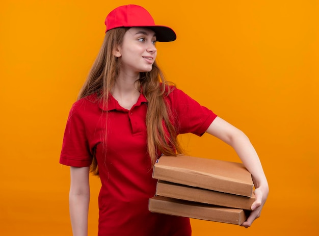 Glimlachend jong leveringsmeisje in rode uniforme holdingspakketten die rechterkant op geïsoleerde oranje ruimte bekijken