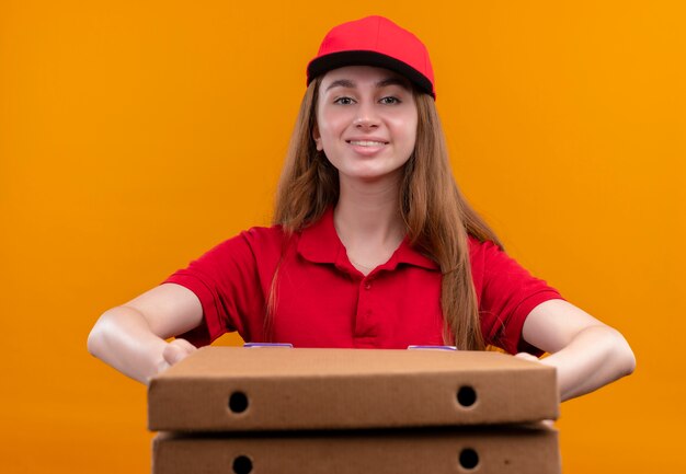 Glimlachend jong leveringsmeisje in rood uniform die pakketten op geïsoleerde oranje muur uitrekken