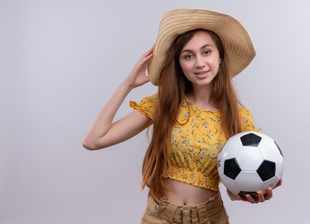 Glimlachend jong meisje dat het voetbalbal van de hoedholding draagt die hand op hoed op geïsoleerde witte muur met exemplaarruimte zet
