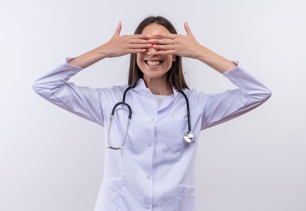 Glimlachend jong meisje dat stethoscoop medische toga draagt behandelde ogen met handen op geïsoleerde witte achtergrond