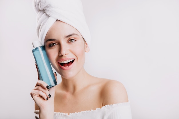 Glimlachend jong meisje in handdoek toont hydraterende tonic in blauwe fles. Portret van groenogig model zonder make-up.