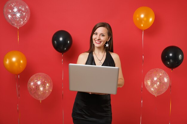 Glimlachend jong meisje in kleine zwarte jurk werken op laptop pc-computer terwijl vieren op rode achtergrond luchtballonnen. internationale vrouwendag happy new year verjaardag mockup party concept vakantie.
