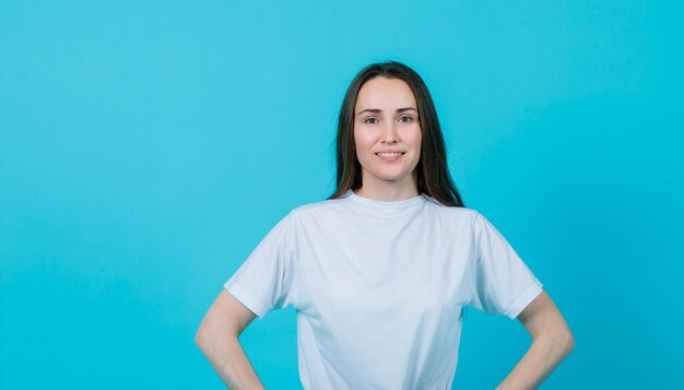 Glimlachend jong meisje kijkt naar de camera door de handen op de taille op een blauwe achtergrond te leggen