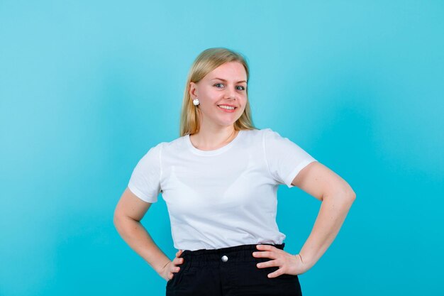 Glimlachend jong meisje kijkt naar de camera door de handen op de taille op een blauwe achtergrond te leggen