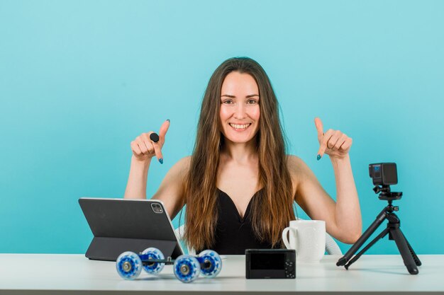Glimlachend jong meisje toont haar camera en tablet met wijsvingers op blauwe achtergrond