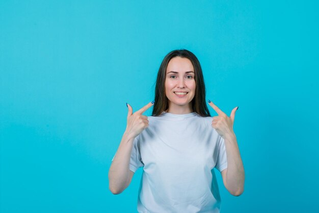 Glimlachend jong meisje toont haar glimlach met forefinegrs op blauwe achtergrond