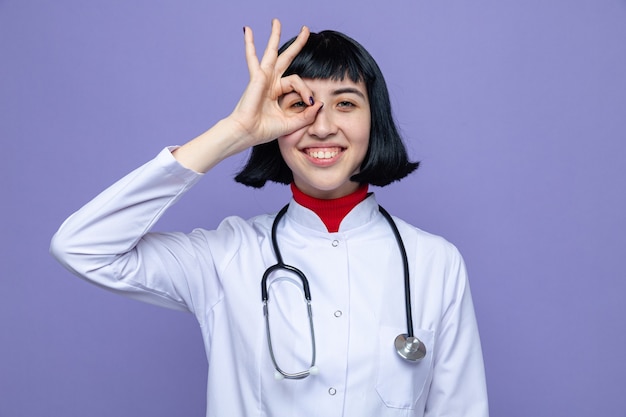 Glimlachend jong mooi Kaukasisch meisje in doktersuniform met stethoscoop kijkend naar de voorkant door vingers