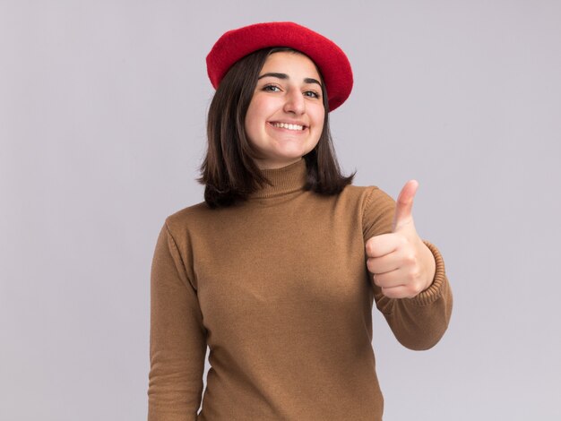 Glimlachend jong mooi Kaukasisch meisje met baret hoed duimen omhoog geïsoleerd op een witte muur met kopie ruimte