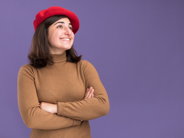 Gratis foto glimlachend jong mooi kaukasisch meisje met baret hoed staande met gekruiste armen kijkend naar kant geïsoleerd op paarse muur met kopieerruimte