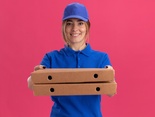 Glimlachend jong mooi leveringsmeisje in de eenvormige dozen van de holdingspizza op roze